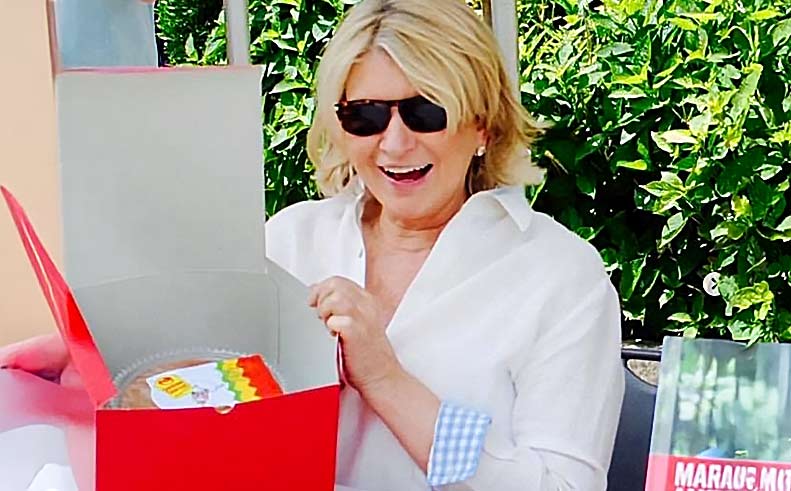 a photo of Martha Stewart wearing sunglasses and a white shirt, holding a red box with a colorful label, outdoors.