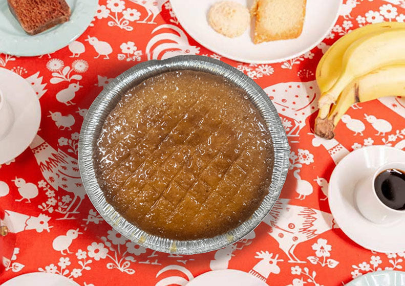 A round aluminum foil pie dish filled with a brown custard-like dessert, placed on a red tablecloth with white animal illustrations. Surrounding props include plates with bread, bananas, and a cup of coffee.