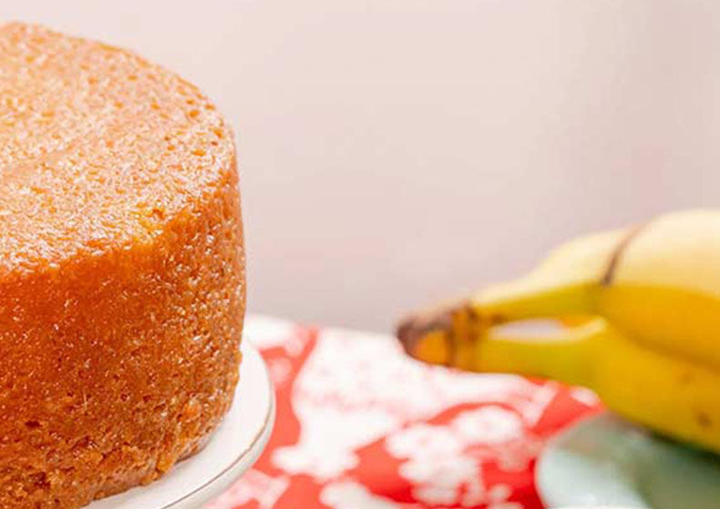 A close-up of a round golden-brown cake on a white plate, with a blurred yellow banana and a red-and-white patterned cloth in the background.