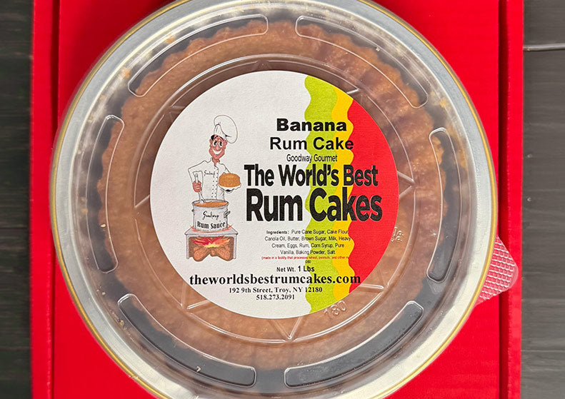 A round clear plastic-lidded container of banana rum cake placed on a red box or sleeve, with a large front label reading 'Banana Rum Cake' and 'The World's Best Rum Cakes' plus an illustrated chef and website text.