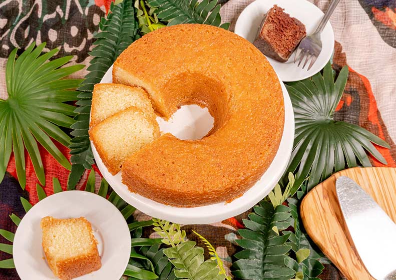 Caribbean rum cake with a slice cut out, surrounded by green leaves and small plates with cake pieces on a colorful background.