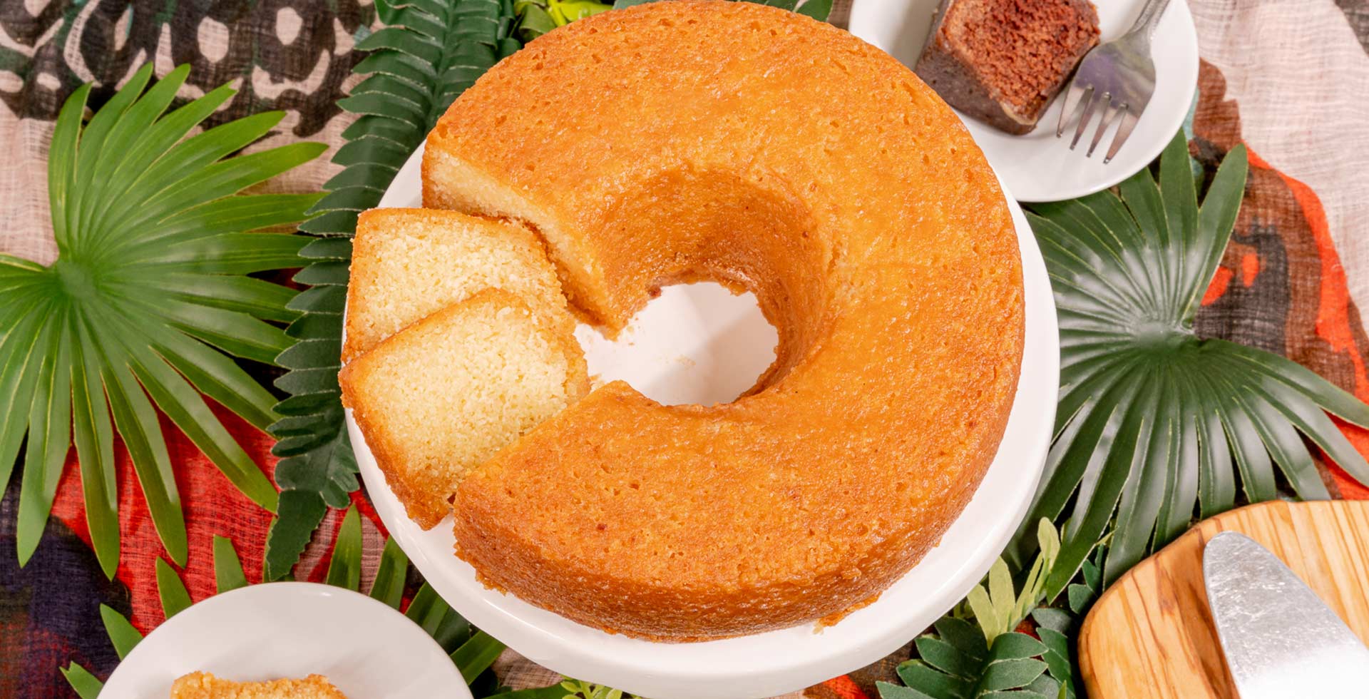 Round cake with a slice cut out on a white plate, surrounded by green leaves.