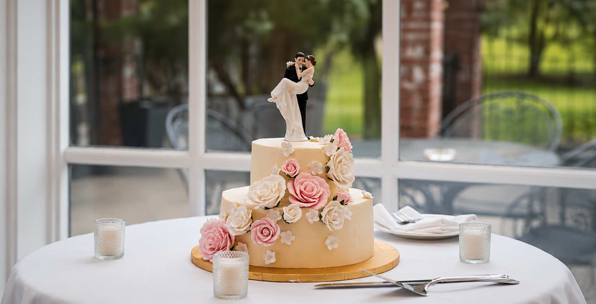 Two-tier wedding cake with decorative flowers and a couple figurine on a table outdoors.