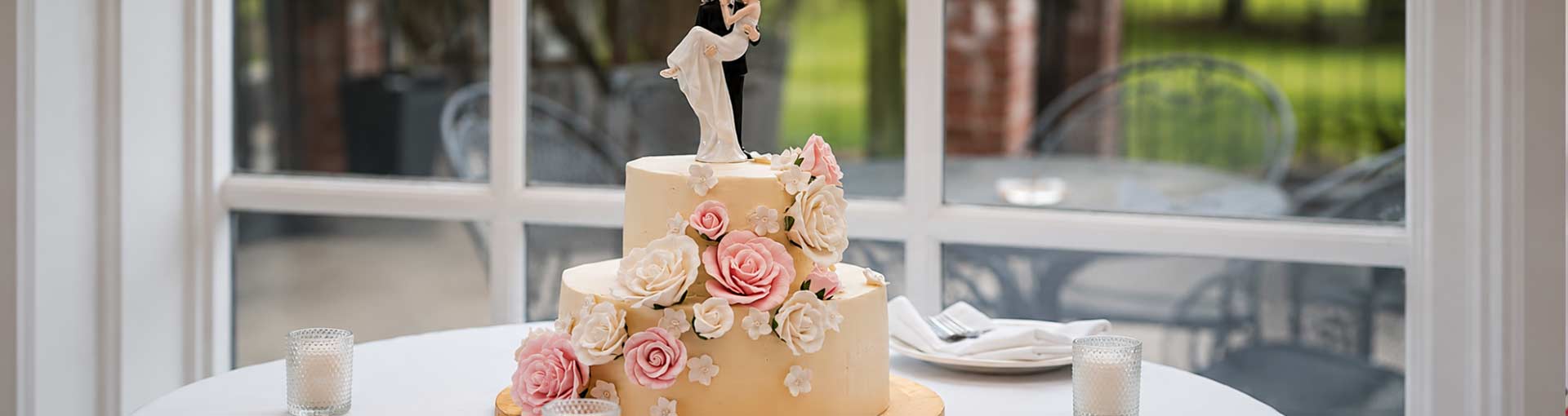 Two-tier wedding cake with decorative flowers and a couple figurine on a table outdoors.