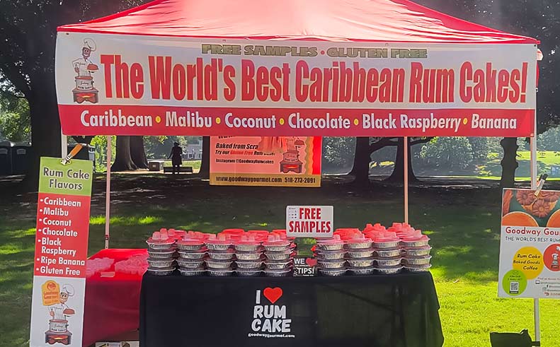 Rum cake stand with promotional signs for Caribbean rum cakes in a park setting.