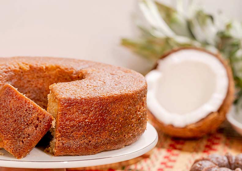 malibu rum cake with a slice cut out on a white plate, coconut halves, and pineapples in the background.