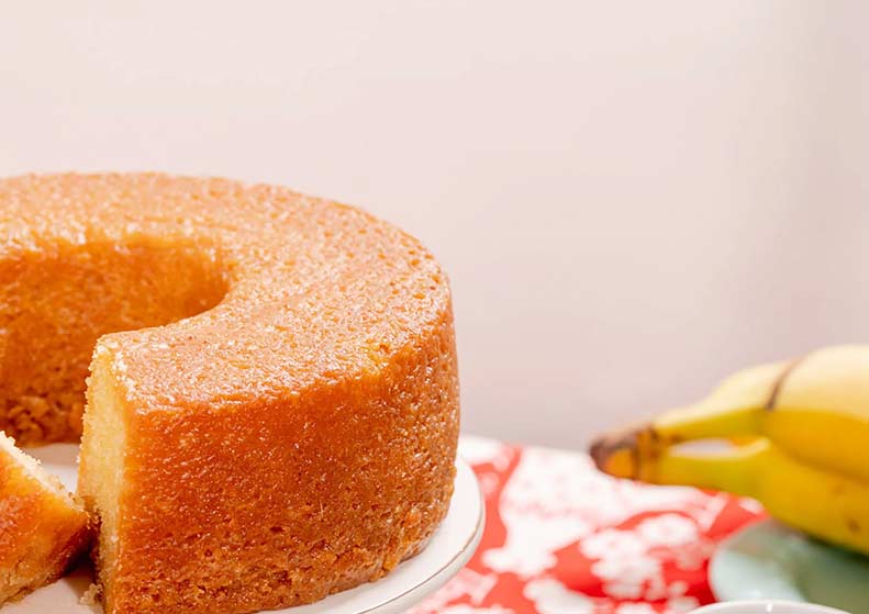 Round cake with a slice cut out on a plate, next to two bananas on a patterned tablecloth.
