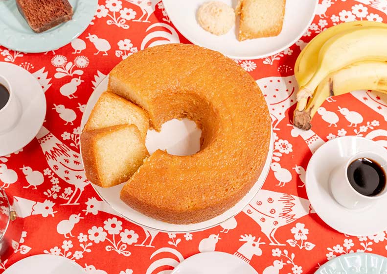 Bundt cake with a slice removed on a decorative tablecloth with coffee and bananas.